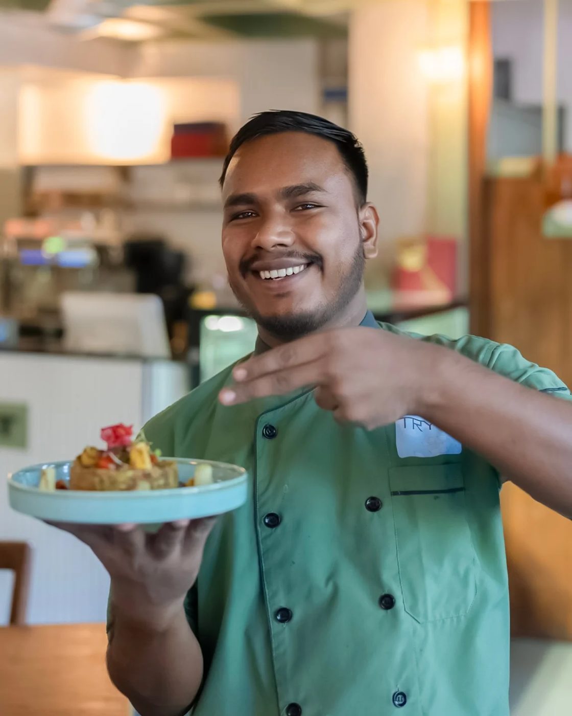 A chef in a green jacket smiling and pointing to a plate he is holding