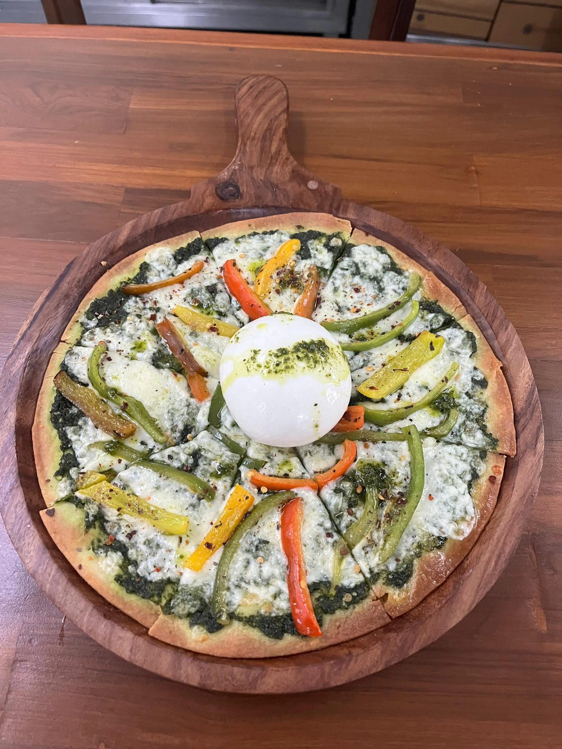 A pizza topped with burrata, peppers and pesto on an olive wood board