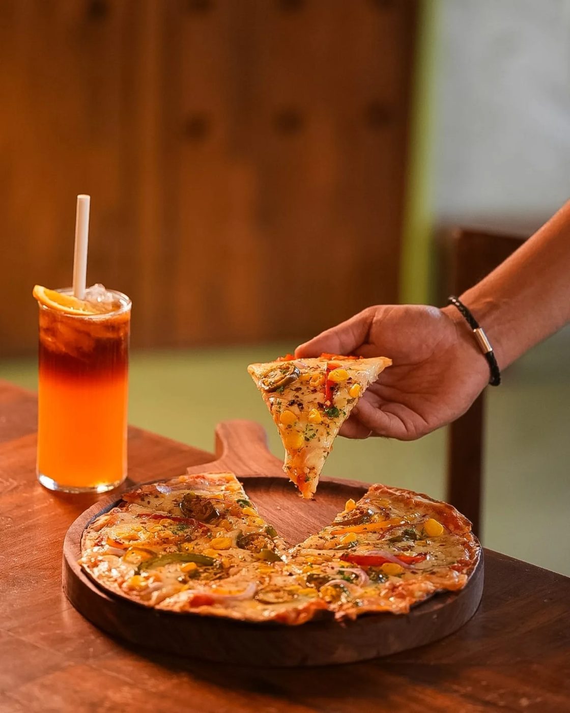 A hand lifting a slice from a thin crust pizza beside an iced tea